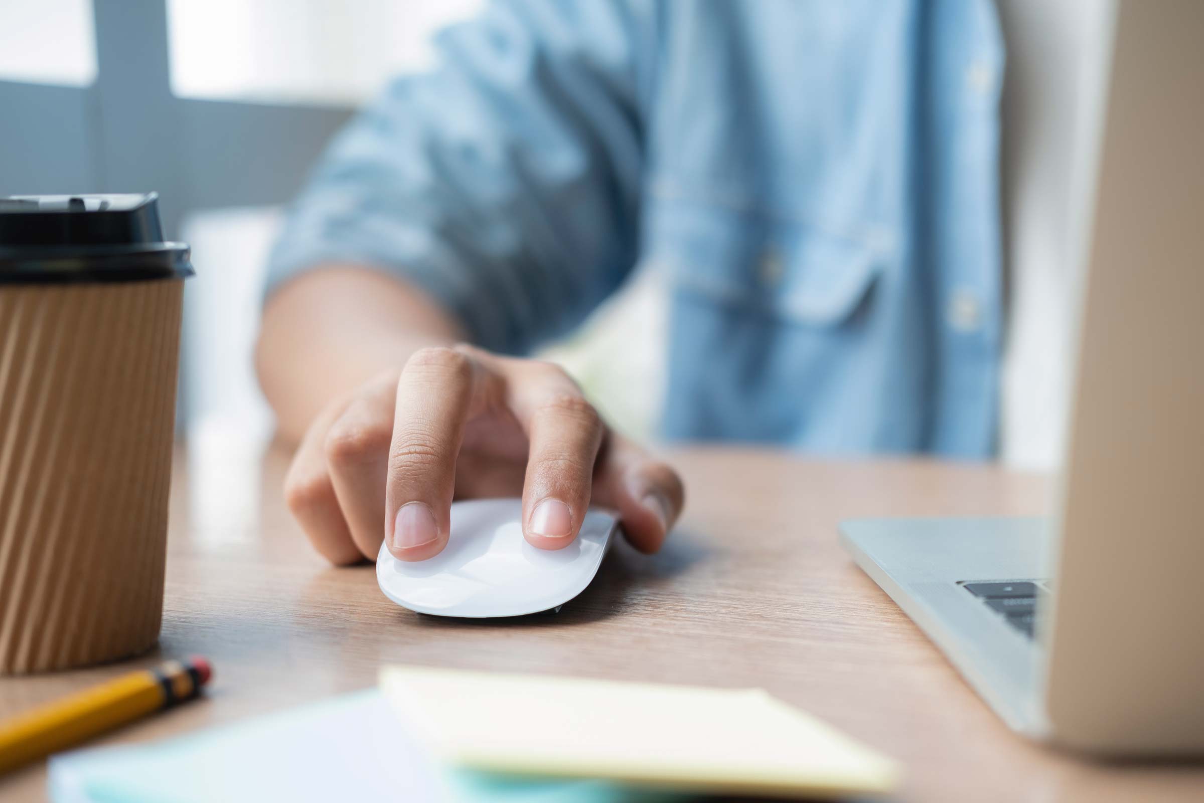 Person using a computer mouse at a desk with a laptop and coffee cup.
